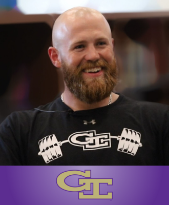 Coach Pat Doyle smiling and sitting in a chair in a school library with the GI logo. 