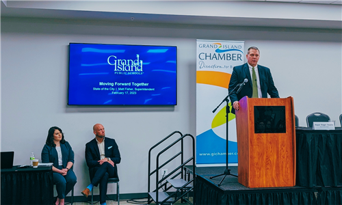 Mr. Matt Fisher at the podium speaking at the State of the City event with two onlookers to the left listening in.